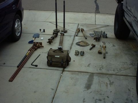 1990 Ford Bronco - Assorted vehicle components and tools are organized on a paved surface, with parts of two vehicles visible at the edges.