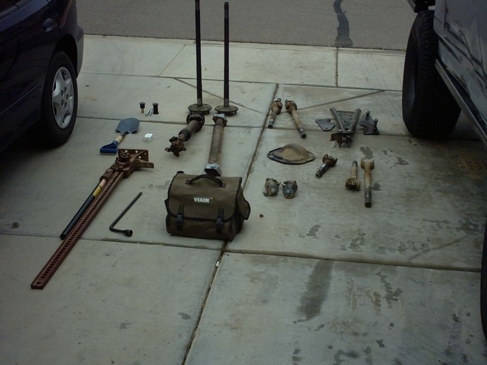 Various vehicle parts including axles, driveshafts, and a Hi-Lift jack on a driveway