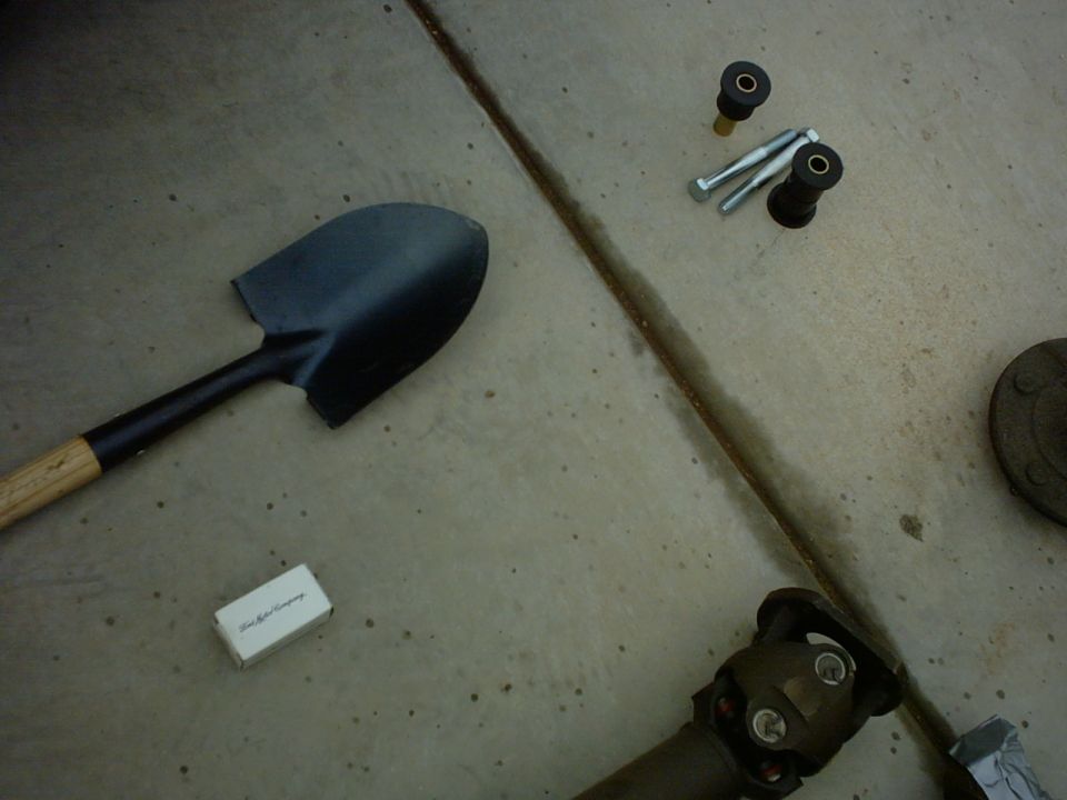 Shovel, bolts, bushings, and a driveshaft U-joint on concrete. Small white box has text