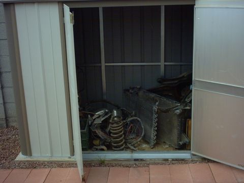 1990 Ford Bronco - Various car parts, such as a radiator and coil spring, are stored inside an open shed.