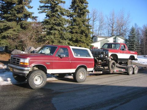 1985 Ford Bronco - A red Ford Bronco tows a red and white Ford pickup truck on a trailer, with trees and houses in the background.