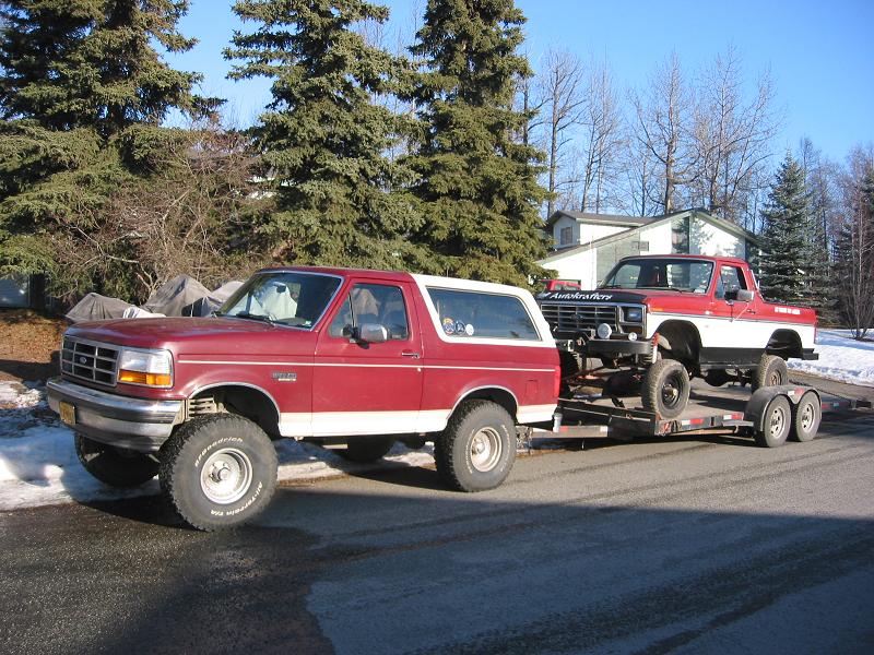 Lifted red and white Ford Bronco towing a trailer with a stripped Ford truck in the snow