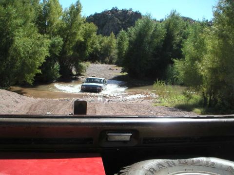 1990 Ford Bronco - A 1990 Ford Bronco navigates a water crossing surrounded by lush greenery and arid hills.