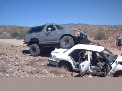 1990 Ford Bronco - A lifted 1990 Ford Bronco is positioned on top of a wrecked white sedan in a dry, arid environment.