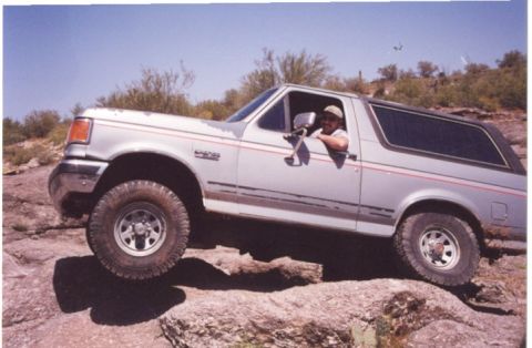 1990 Ford Bronco - Driver navigates a 1990 Ford Bronco over a rocky trail under a clear blue sky.