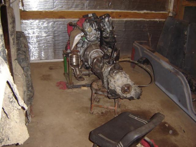 Engine and transmission assembly on a stand, next to a tire and part of a vehicle body