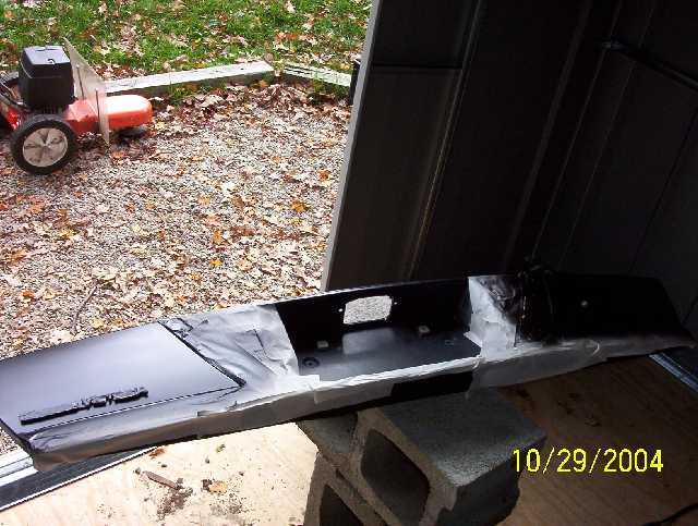 Black car bumper on cinder blocks, partially covered in paper. Lawn mower in background