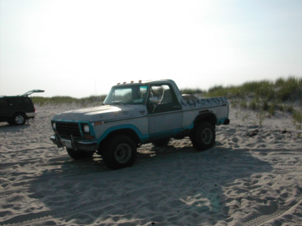 White and teal lifted Ford 4x4 on a sandy beach with dunes behind it
