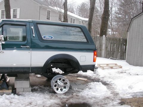 1994 Ford Bronco - Side view of a Ford Bronco, lifted on blocks with a wheel off, in a snowy yard.