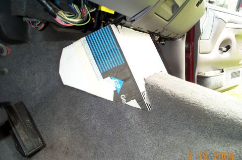 1996 Ford Bronco - Taped-together cardboard mockup of a wedge-shaped under-dash enclosure sitting on the gray carpet of a Ford truck footwell, with the dash p…