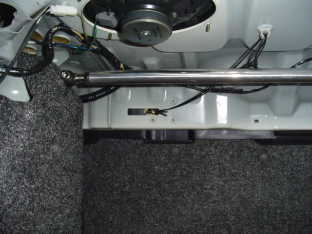 Chrome power antenna mast resting in trunk area with rear speaker and wiring visible above gray carpet