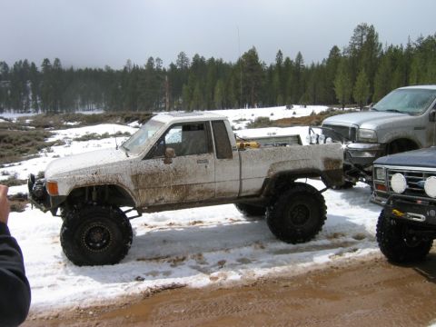 1985 Ford Bronco - A lifted white pickup truck is parked in a snowy, muddy area with trees in the background.