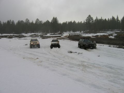1985 Ford Bronco - Three SUVs navigate a snowy terrain under an overcast sky, with a line of trees in the distance.