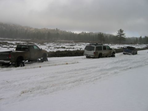 1985 Ford Bronco - Three vehicles are stuck in a snowy landscape with a forest and hills visible in the distance.