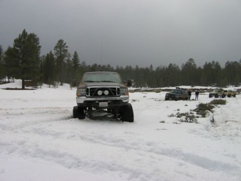 1985 Ford Bronco - A lifted pickup truck navigates a snowy landscape with other vehicles and people visible in the distance.