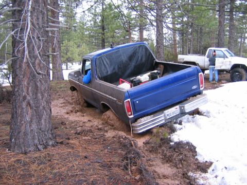 1985 Ford Bronco - A blue Ford Bronco is stuck in the mud, with a white pickup truck visible in the background.