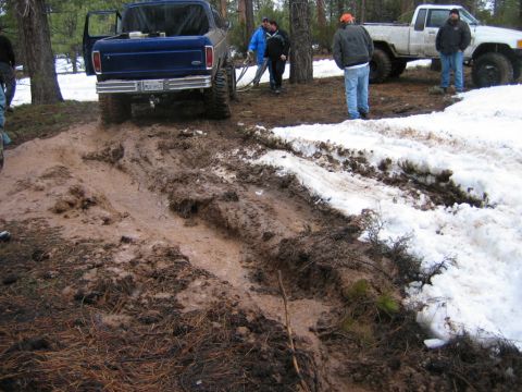 1985 Ford Bronco - A blue Ford Bronco struggles in a muddy terrain with a white Ford pickup truck in the background.