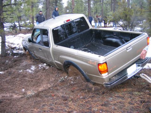 1985 Ford Bronco - A gold pickup truck with a 4x4 badge is stuck in the mud with several people watching.