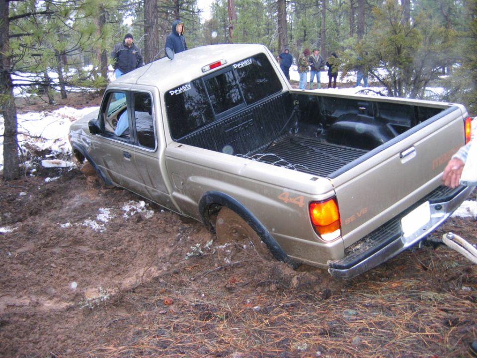 Gold Mazda 4x4 pickup stuck in mud with people watching in snowy woods