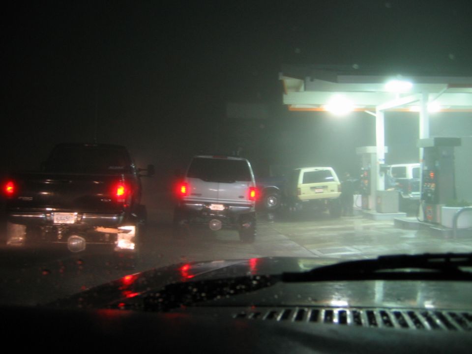 Foggy night gas station with several vehicles seen through a rain-covered windshield.