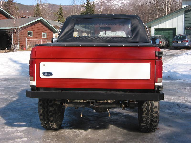 Rear of red lifted Ford truck with soft top, tube bumper, and big mud tires on snow