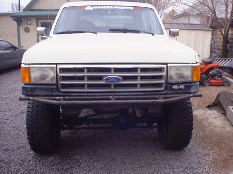 1988 Ford Bronco - Head-on front view of a lifted white Ford 4x4 on a gravel driveway, showing the bar-style grille with blue Ford oval, amber corner lenses,…