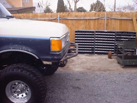 1988 Ford Bronco - Three-quarter front view of a lifted two-tone white-over-blue Ford truck in a gravel yard, wearing a bare steel tube front bumper, mud-terr…