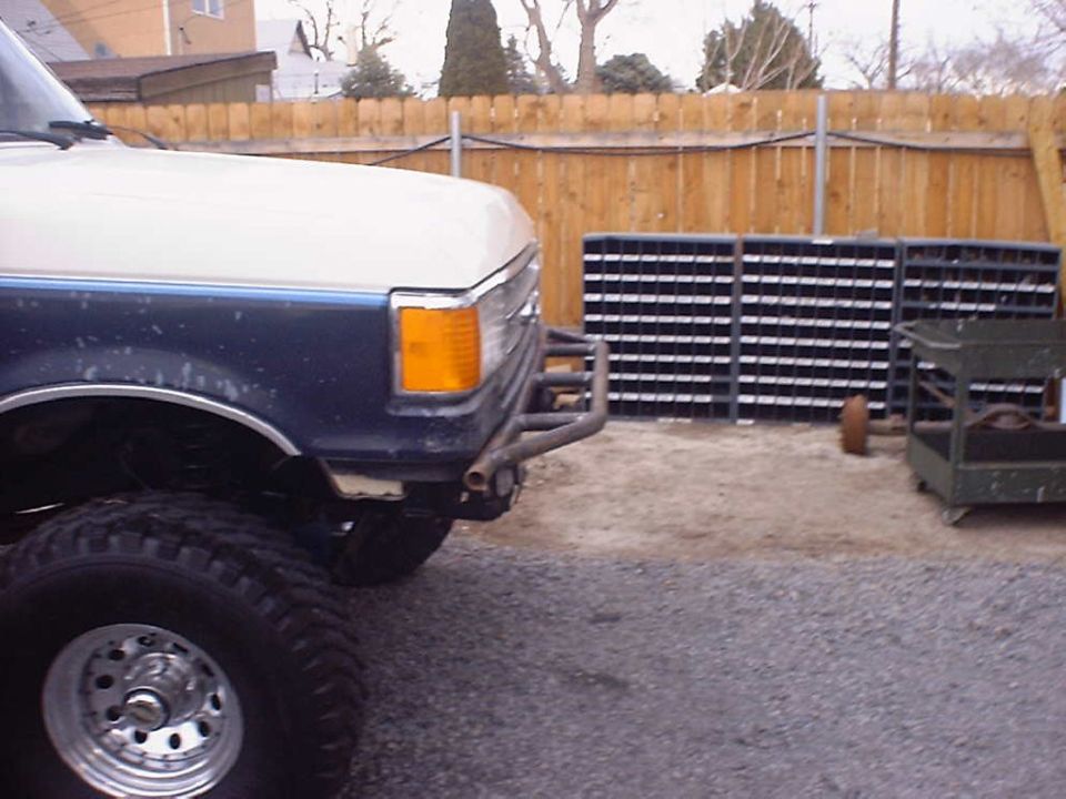 Lifted two-tone white and blue Ford truck with tube bumper and mud tires in a gravel yard