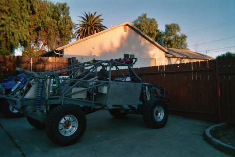 1990 Ford Bronco - bare tube-chassis off-road race buggy with coilover shocks sits in a backyard beside a fence at golden hour.