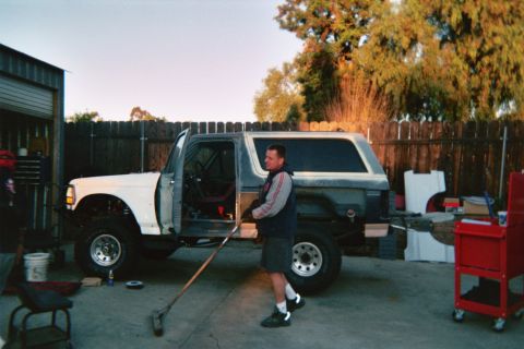 1990 Ford Bronco - man stands beside a white full-size Ford Bronco with its door open in a backyard workshop at sunset, broom in hand.