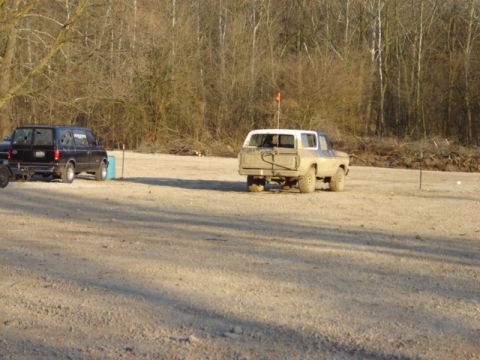 1992 Ford Bronco - An older model SUV and a dark van are parked in an open, dusty area with a wooded background.