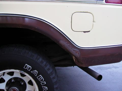 1979 Ford Bronco - Close-up of the left rear quarter panel of a cream and dark red two-tone truck, showing heavy rust bubbling and perforation along the lower…
