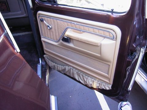 1979 Ford Bronco - Open passenger door of a dark maroon truck showing a tan door panel with quilted vinyl upper, chrome trim strip, molded armrest with recess…