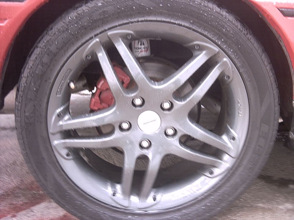 Close-up of a car's front wheel with a red brake caliper. The wheel has a multi-spoke design and 'ITALY' is visible on the tire sidewall