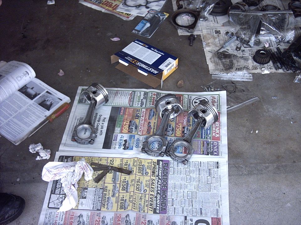 Engine pistons and connecting rods laid out on newspapers, with a manual and parts box nearby