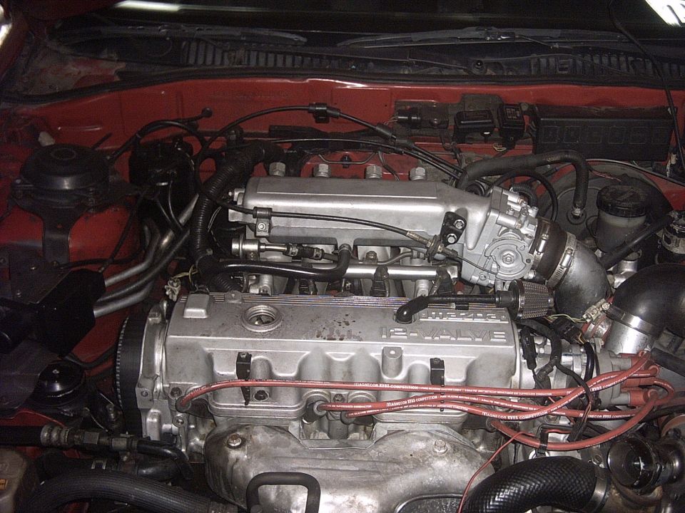 Engine bay with a red car exterior visible, showing a 16-valve engine with silver intake manifold and red spark plug wires