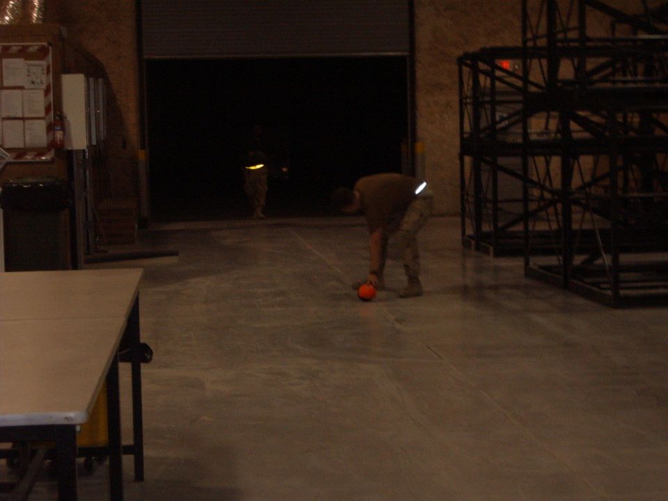 person picking up an orange ball in a warehouse, another person stands in doorway