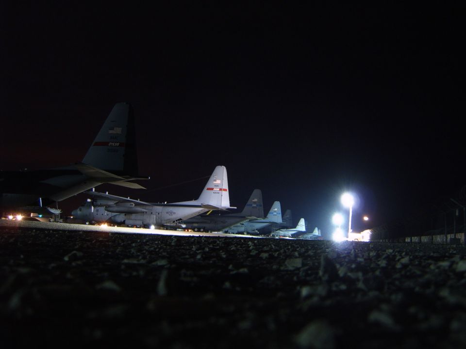 Several military transport planes parked on a gravel tarmac at night under bright lights
