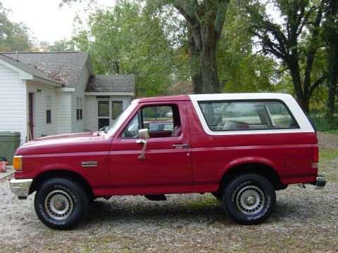 1988 Ford Bronco - This red Ford Bronco with a white top is seen from the side, parked on a gravel surface next to a house.