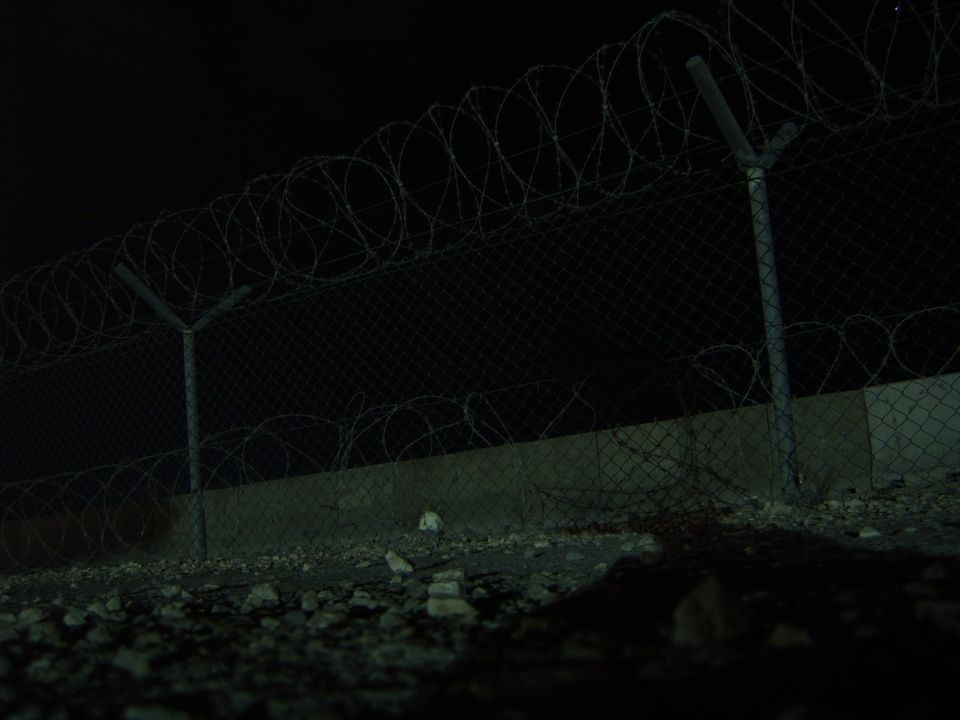 Barbed wire fence and concrete barrier at night, with gravel on the ground