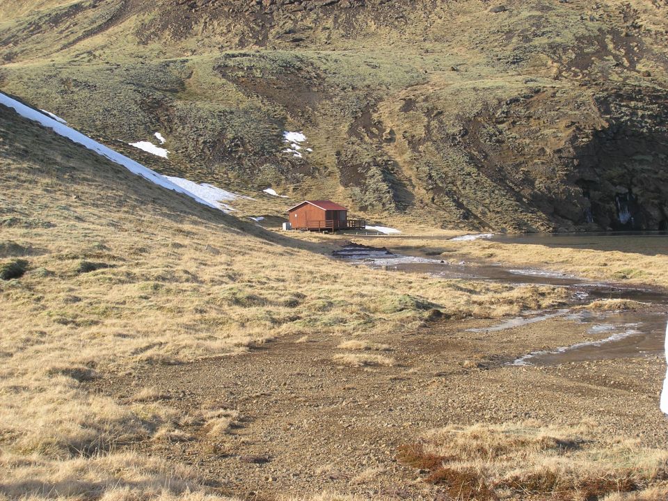 Small wooden cabin with a red roof sits in a valley with dry grass, snow patches, and a stream