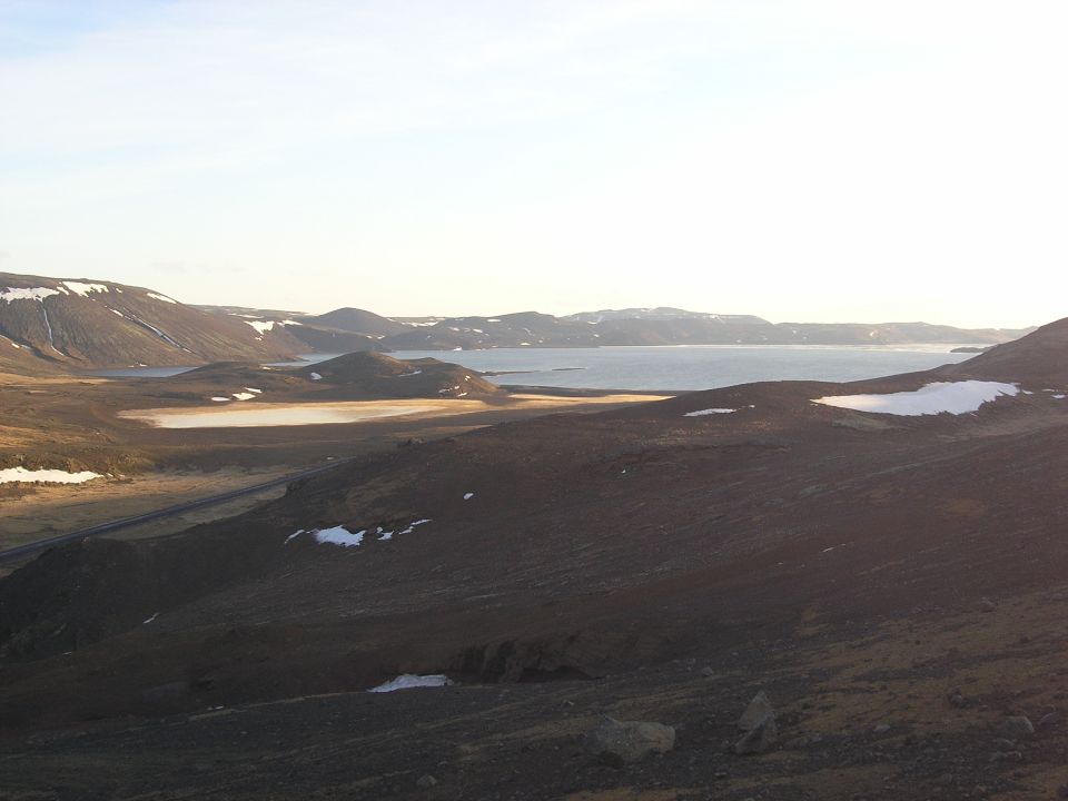 Vast Icelandic landscape with mountains, a lake, and snow patches under a bright sky