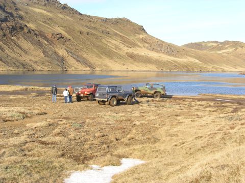 2001 Jeep Wrangler - A group of off-road vehicles are parked near a body of water, with a large mountain range visible behind them.