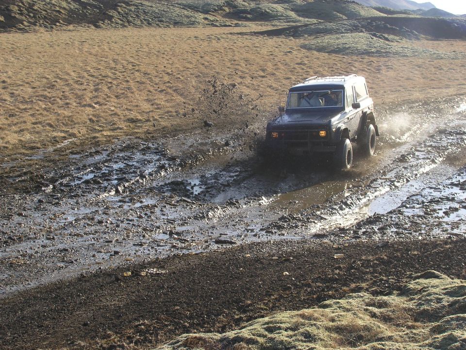 Dark SUV driving through mud, splashing water. Visible text 'SUPER MOTORS'