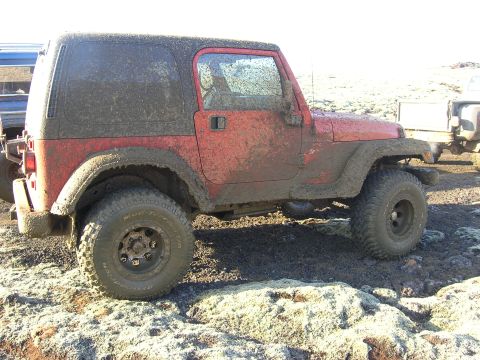 2001 Jeep Wrangler - A muddy red 2001 Jeep TJ is parked off-road, with another vehicle visible to the left and a trailer to the right.