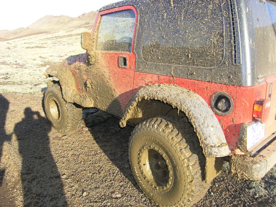 Mud-covered red 2001 Jeep TJ parked on a dirt road with mountains in the background