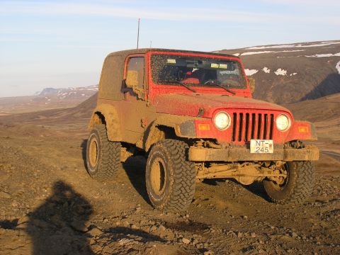 2001 Jeep Wrangler - A muddy red 2001 Jeep TJ navigates a rough, off-road terrain under a clear sky.