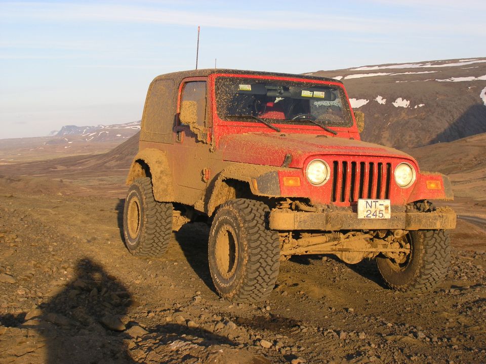 Red 2001 Jeep TJ covered in mud, parked on a dirt road with mountains in the background