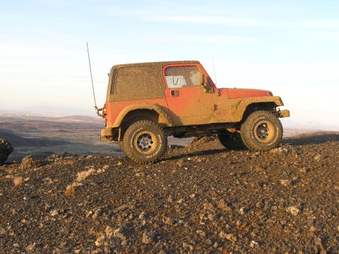 2001 Jeep Wrangler - A muddy red 2001 Jeep TJ is parked on a rocky incline, with a vast landscape visible in the background.