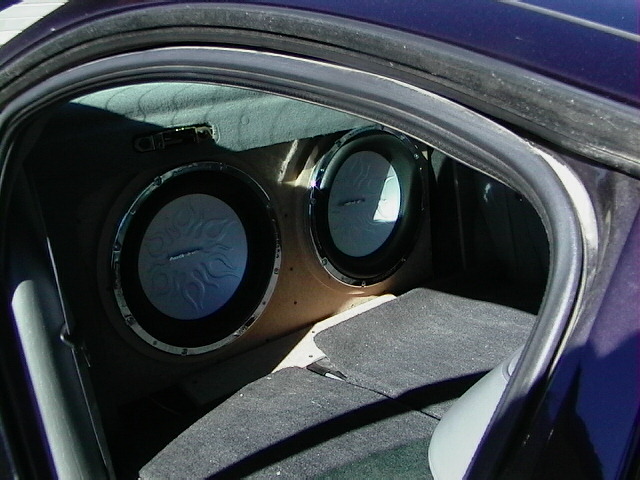 Two flame-cone subwoofers in a custom box behind rear seats of a dark blue car